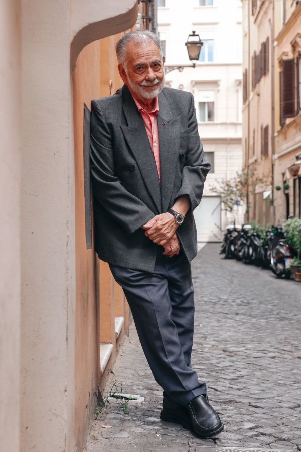 Un uomo anziano con capelli e barba grigi si appoggia con disinvoltura a un muro su una strada acciottolata, vestito con un blazer grigio, una camicia rossa e pantaloni scuri. Sullo sfondo, moto e alti edifici costeggiano la strada.