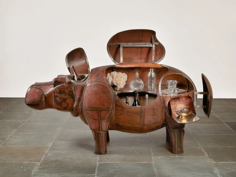 Un ippopotamo in bronzo scolpito con diversi scomparti aperti, che rivelano scaffali pieni di bicchieri e bottiglie, si trova su un pavimento piastrellato su uno sfondo semplice.