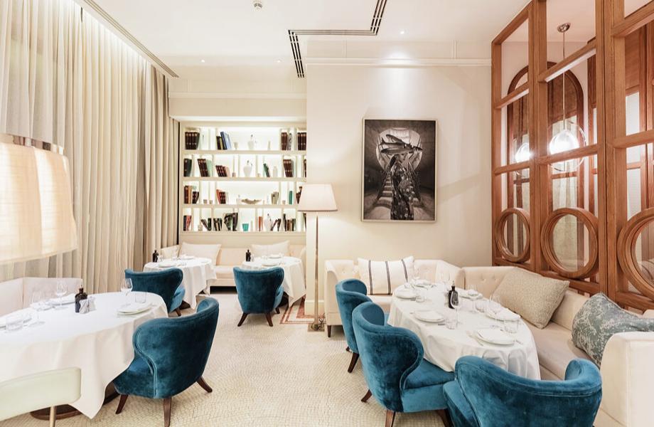 Elegant restaurant interior with round tables, white tablecloths, blue velvet chairs, beige sofas, soft lighting, bookshelves, and framed artwork on the wall, creating a cozy and sophisticated atmosphere.