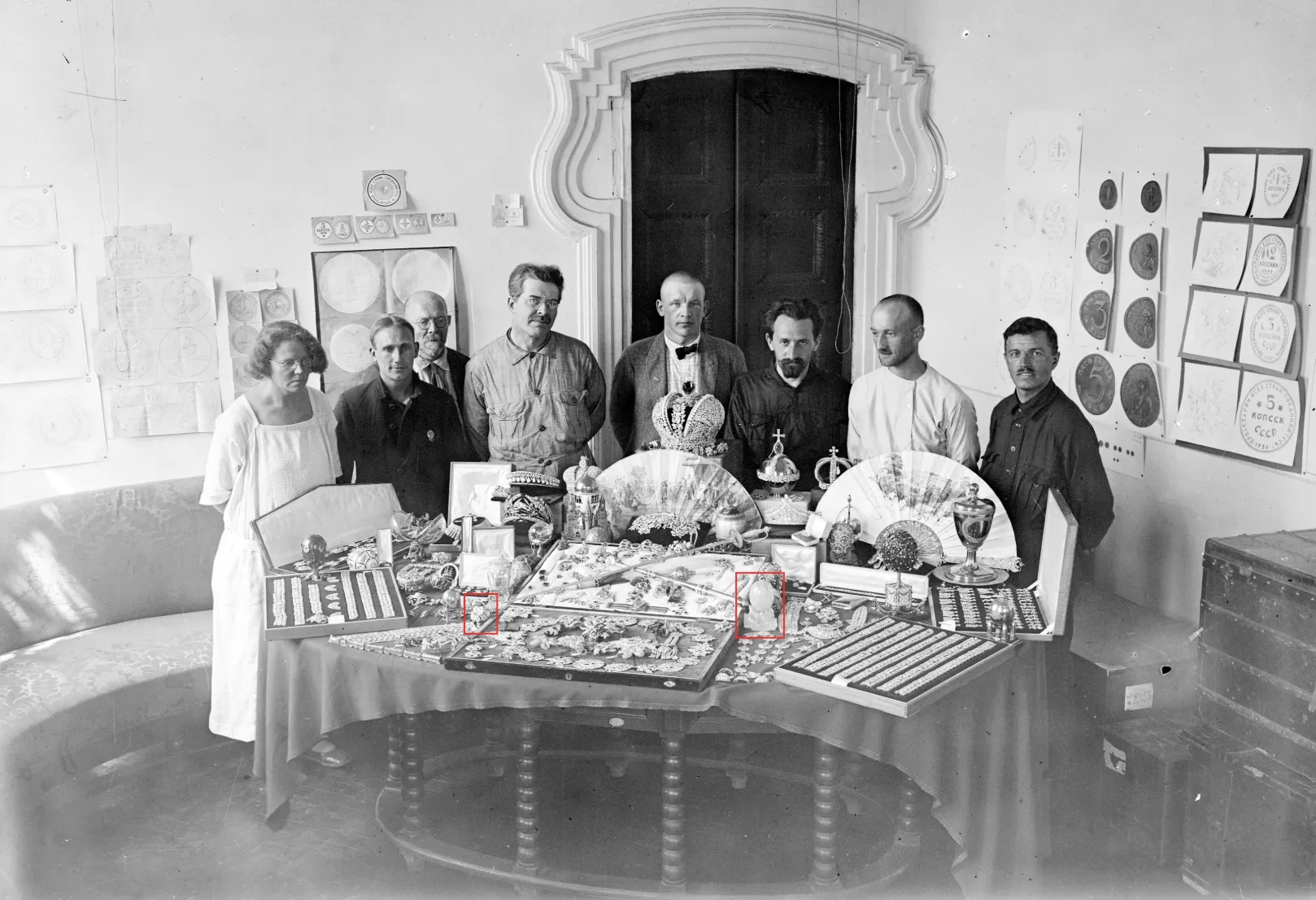 Un gruppo di otto persone, uomini e donne, si trova dietro un tavolo coperto di oggetti decorativi, ventagli, pezzi ispirati a Fabergé e opere d'arte. Alle pareti della stanza sono esposti disegni e opere d'arte incorniciate.