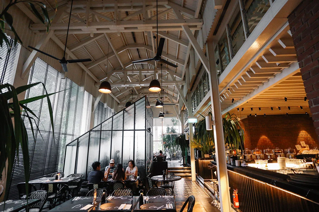 A modern, stylish restaurant interior with high ceilings, exposed beams, hanging lights, and large windows. Several people sit at tables, and plants decorate the space, creating a bright, inviting atmosphere.