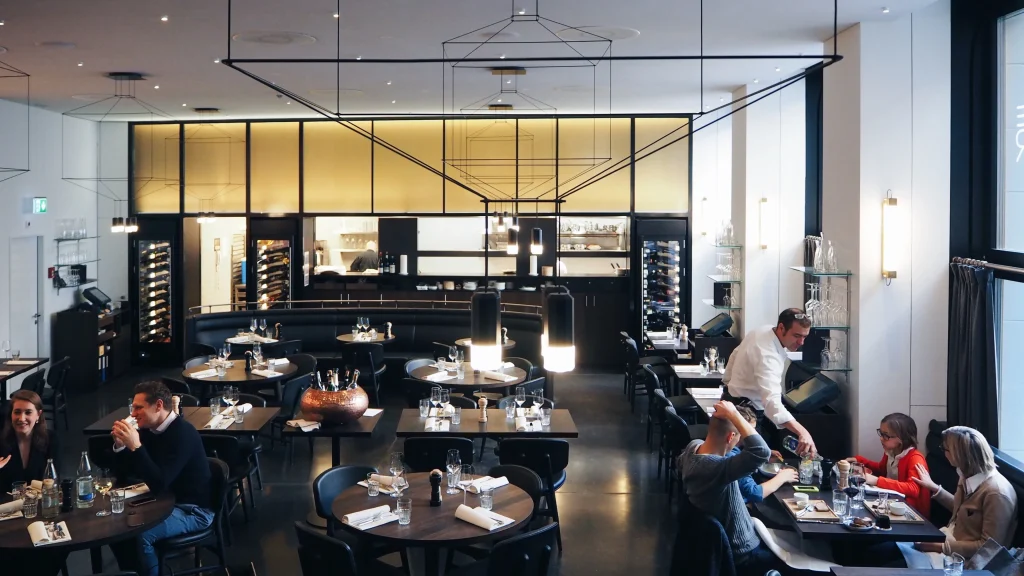 A modern restaurant interior with several groups of people dining at round tables. A waiter is serving a table, and large windows let in natural light. The atmosphere appears lively and welcoming.