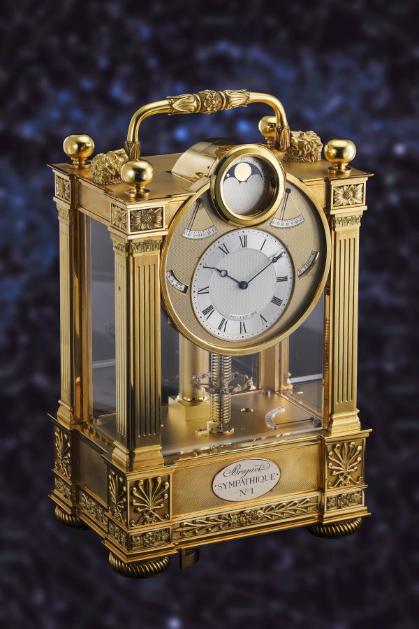 Lussuoso orologio da carrozza in oro con dettagli ornamentali, lati in vetro trasparente, numeri romani sul quadrante bianco e una maniglia decorativa sulla parte superiore, su uno sfondo scuro e sfocato.