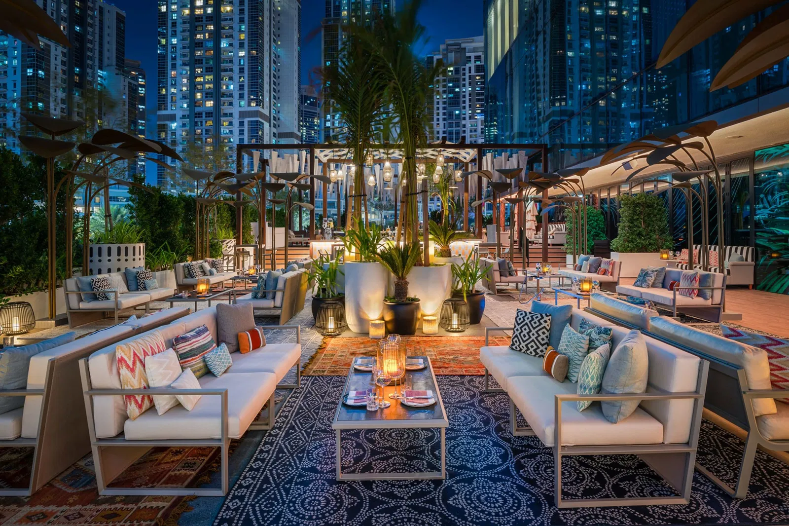Stylish outdoor lounge at night with cozy sofas, colorful pillows, patterned rugs, lit candles, and lush plants, set against the backdrop of illuminated city skyscrapers.