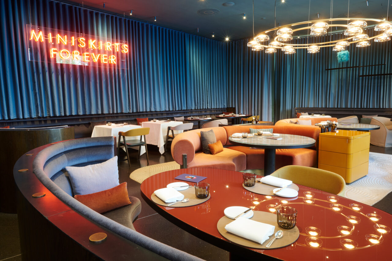 Interno di un ristorante moderno a Lugano con sedute curve e colorate, tavoli rotondi per mangiare, un'insegna al neon "MINISKIRTS FOREVER" su una parete blu plissettata e un lampadario circolare con luci a globo in alto.