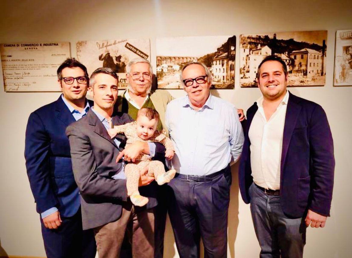 Sei persone, tra cui un bambino, posano in interni davanti a foto d'epoca. Cinque adulti in abiti business-casual sorridono all'obiettivo; un uomo tiene in braccio il bambino. La calda scena riflette lo spirito familiare del Biscottificio Grondona.