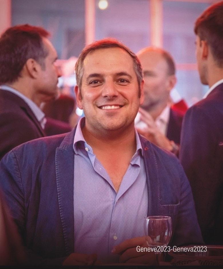 Un uomo sorridente con un blazer blu siede a un tavolo con un bicchiere di vino in mano, con i prodotti del Biscottificio Grondona nelle vicinanze e altre persone impegnate in una conversazione sullo sfondo sfocato di un evento sociale.