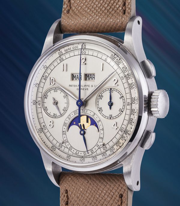 Primo piano di un orologio da polso di lusso Patek Philippe con cinturino in pelle beige, cassa argentata, quadranti multipli e indicazione delle fasi lunari su uno sfondo blu sfumato.