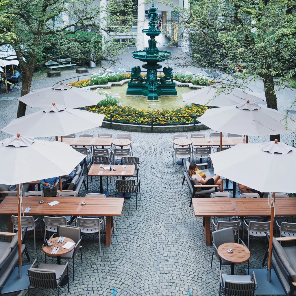 Outdoor café with wooden tables and white umbrellas set on a cobblestone patio. A few people are seated. In the background, there is a circular fountain surrounded by flowers and trees.