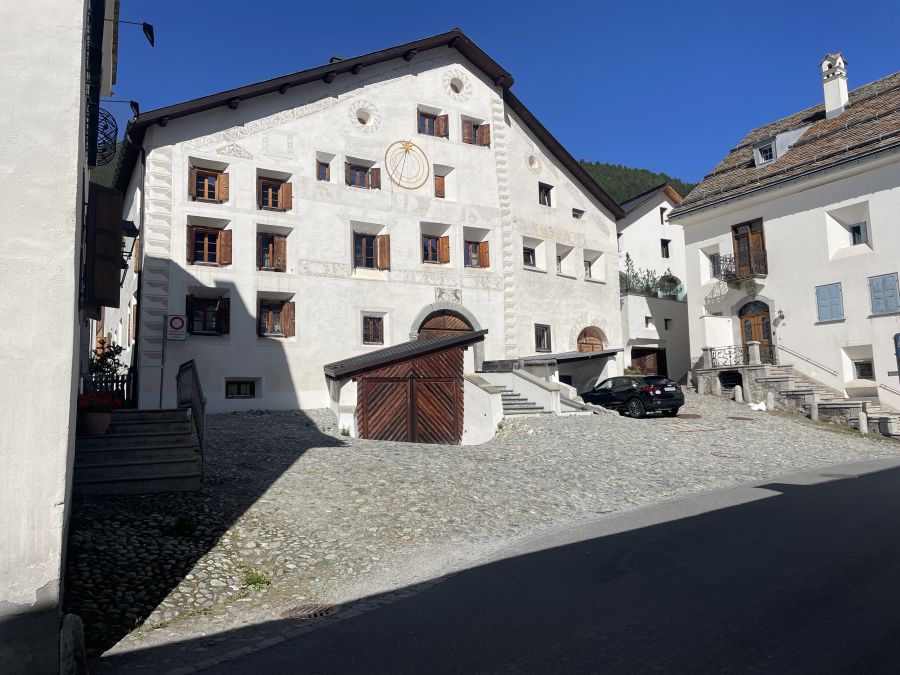 Un grande edificio bianco con persiane in legno e un disegno circolare in engadina arte sul muro si trova in un cortile soleggiato e acciottolato, con una piccola baracca in legno e due auto parcheggiate nelle vicinanze. Il cielo azzurro e altri edifici bianchi sono visibili.