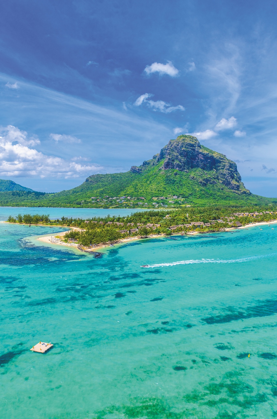 Vista aerea di una montagna verde e lussureggiante sopra una costa tropicale, dove acque turchesi e spiagge sabbiose invitano tutti gli amanti della spiaggia. Alberi sparsi incorniciano la riva, mentre una piccola barca sfreccia sul mare sotto un cielo luminoso e parzialmente nuvoloso.