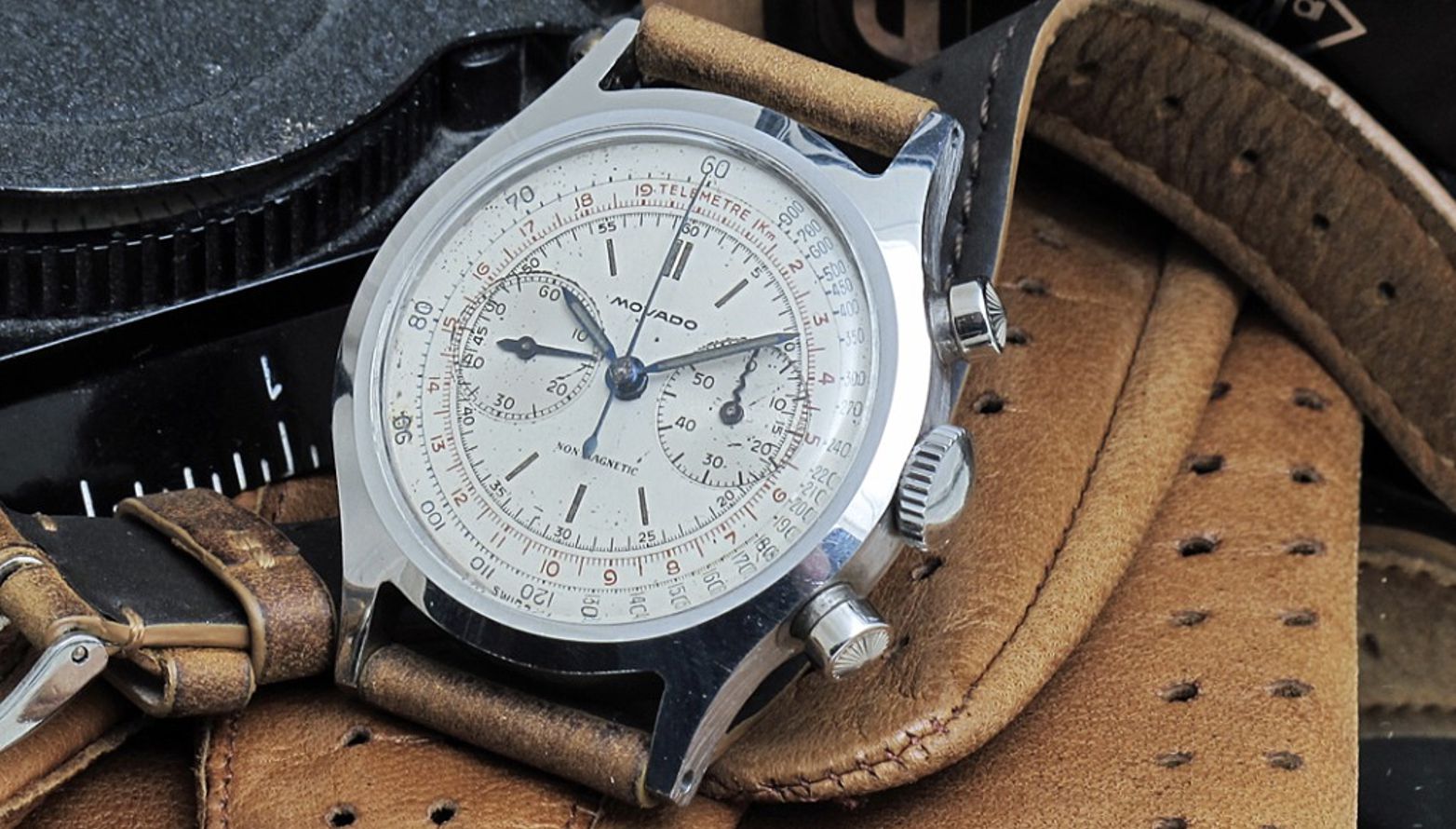 Un orologio da polso cronografo Movado vintage con quadrante color crema, indici neri e due contatori, si trova su un cinturino in pelle marrone e un guanto in pelle marrone, accanto a uno strumento nero.