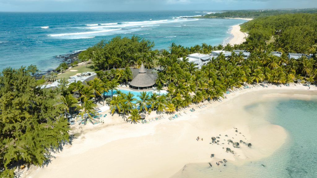Veduta aerea di un resort sulla spiaggia tropicale con sabbia bianca, acqua turchese, fitte palme e un edificio centrale con tetto di paglia circondato da sedie a sdraio e ombrelloni lungo il litorale.