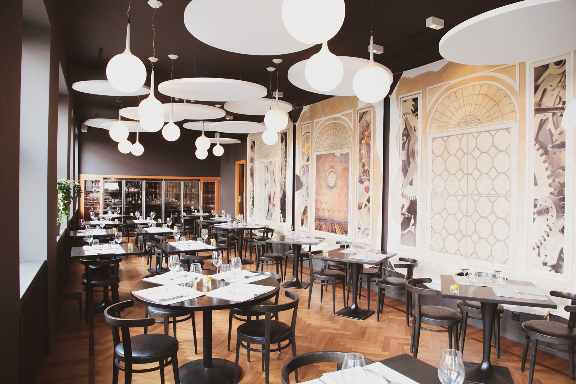 Interno di un ristorante moderno ispirato a Reggio Emilia, con tavoli e sedie neri, pavimenti in parquet di legno, murales artistici, luci a sospensione a globo e una vetrina per i vini in vetro. I tavoli sono elegantemente apparecchiati con bicchieri e tovaglioli.