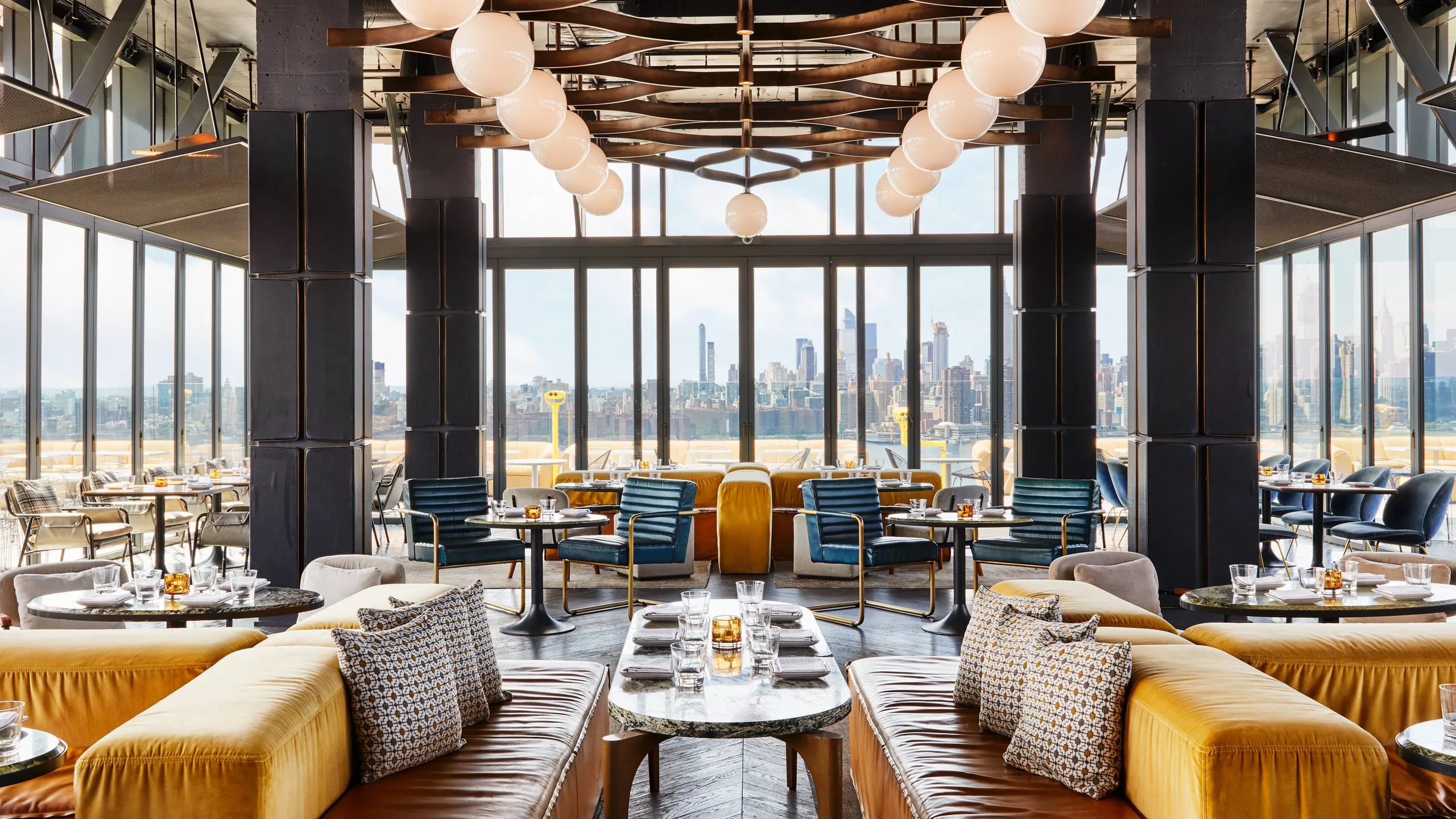 Interno di un ristorante moderno e raffinato con vetrate a tutta altezza, sedute gialle e blu, tavoli in marmo e luci a sospensione a globo. Le finestre offrono una vista sullo skyline della città sullo sfondo.