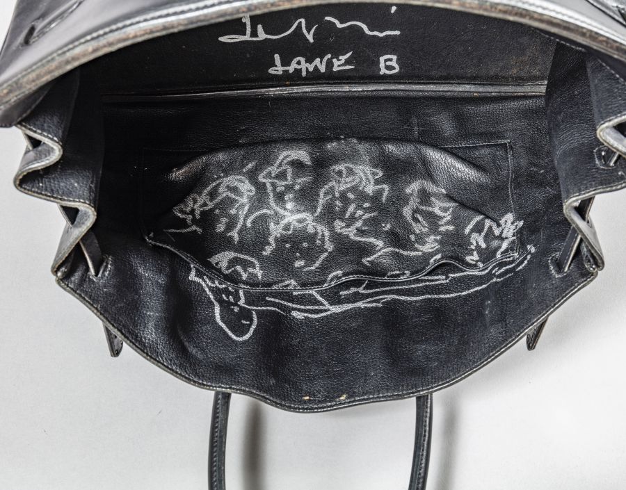 Una borsa di pelle nera aperta che rivela una tasca interna con uno schizzo bianco di diversi volti e figure da cartone animato disegnati sul tessuto nero.