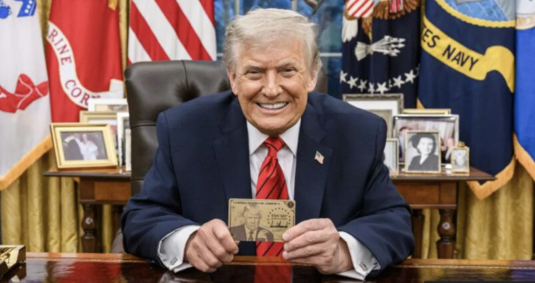Un uomo sorridente in abito scuro e cravatta rossa siede a una scrivania con in mano un biglietto d'oro di Trump con la sua immagine. Dietro di lui ci sono bandiere statunitensi e foto incorniciate, a suggerire che l'ambiente è un ufficio formale.
