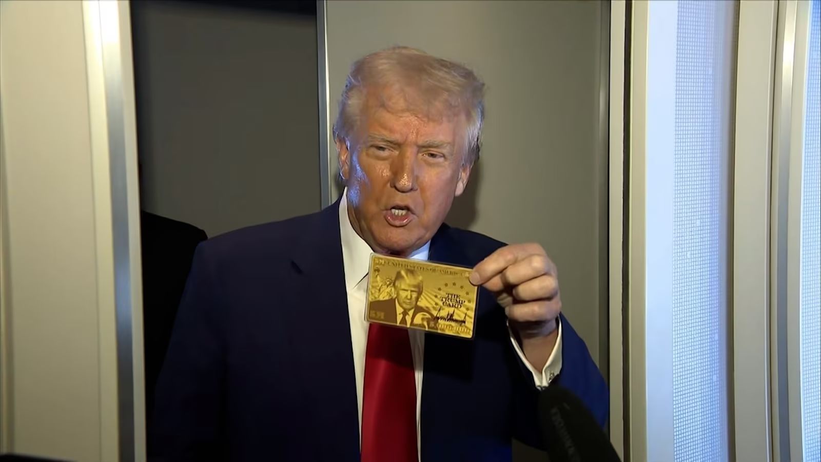 Un uomo in abito e cravatta rossa tiene in mano un biglietto d'oro di Trump, in piedi davanti a una struttura metallica o a una porta, mentre parla o fa un'espressione.