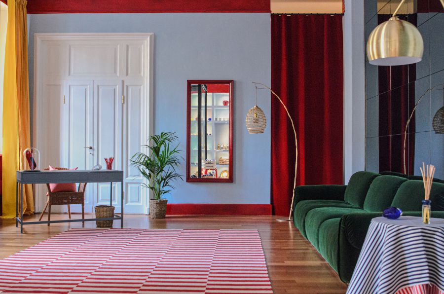 Un soggiorno elegante con divano verde, tappeto a righe bianche e rosse, pareti blu, luci a sospensione in oro e rattan, scrivania nera con sedia, piante in vaso e tende rosse e oro ispirate a ViBi Venezia. Uno specchio è appeso alla parete.