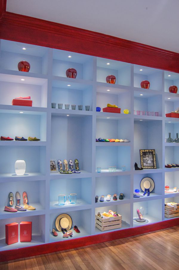Scaffali con cornici rosse espongono varie scarpe colorate, cappelli, pantofole ViBi Venezia, cristalleria, vasi e oggetti decorativi, tutti ordinatamente disposti in singoli scomparti su uno sfondo azzurro.