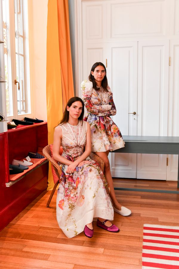 Due donne in una stanza elegante, entrambe con abiti a stampa floreale. Una è seduta su una sedia con scarpe rosa ViBi Venezia; l'altra è in piedi accanto a lei con le braccia incrociate e indossa scarpe bianche. La stanza ha pavimenti in legno, una porta bianca e decorazioni colorate.