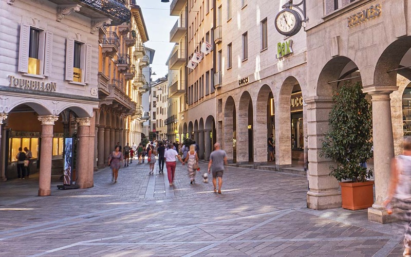 Una pittoresca strada pedonale di Reggio Emilia, fiancheggiata da negozi e passaggi ad arco, dove la gente passeggia e fa acquisti sotto il cielo soleggiato. Le vetrine presentano marchi di lusso e un orologio è montato su uno degli edifici storici.