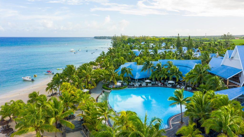 Veduta aerea di un resort sulla spiaggia con una grande piscina circondata da palme, edifici dai tetti blu e acqua limpida e turchese dell'oceano con barche vicino alla riva sabbiosa.