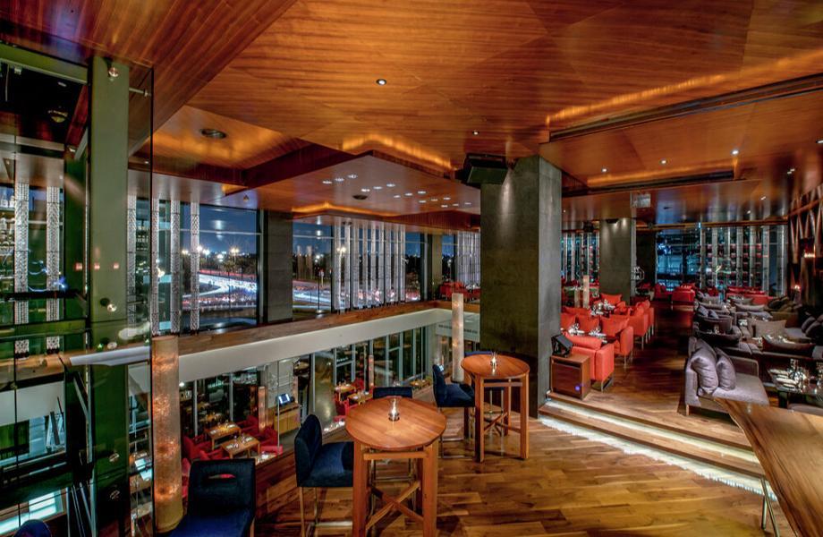 A modern, upscale restaurant interior with wooden floors and ceiling, glass walls, red cushioned seats, and small tables. City lights are visible through the large windows at night.