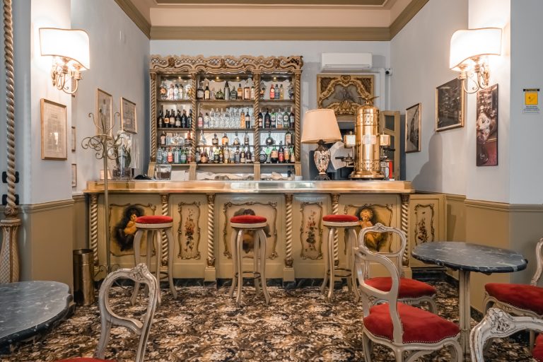 Elegante bar trapanese in stile vintage con dettagli dorati, sgabelli in velluto rosso, tavoli in marmo e un fornitissimo scaffale di liquori dietro il bancone. L'arredamento comprende opere d'arte incorniciate e lampadari classici.
