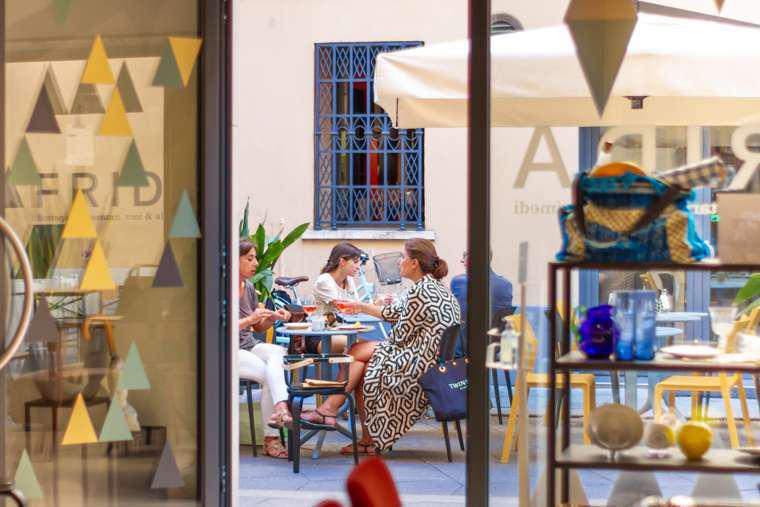 Quattro donne sedute a un tavolo all'aperto a Trapani, mangiano e conversano in un bar. La scena è vista attraverso una porta di vetro con motivi a triangolo, con scaffali di piatti visibili all'interno e un ombrellone che ombreggia l'area di seduta.