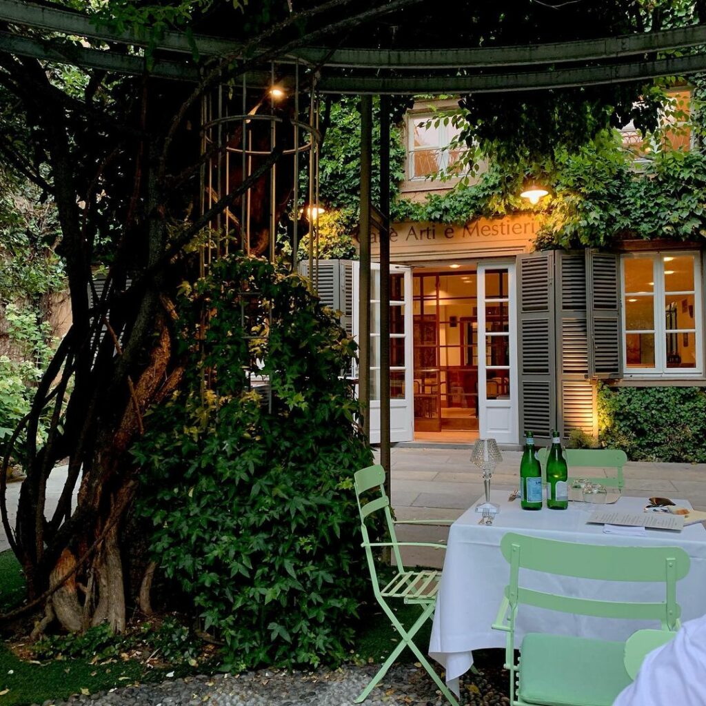 Il patio di un ristorante all'aperto a Trapani con sedie e tavoli verdi per mangiare, circondato da una vegetazione lussureggiante, viti e un edificio illuminato in modo soffuso con porte e finestre aperte sullo sfondo.