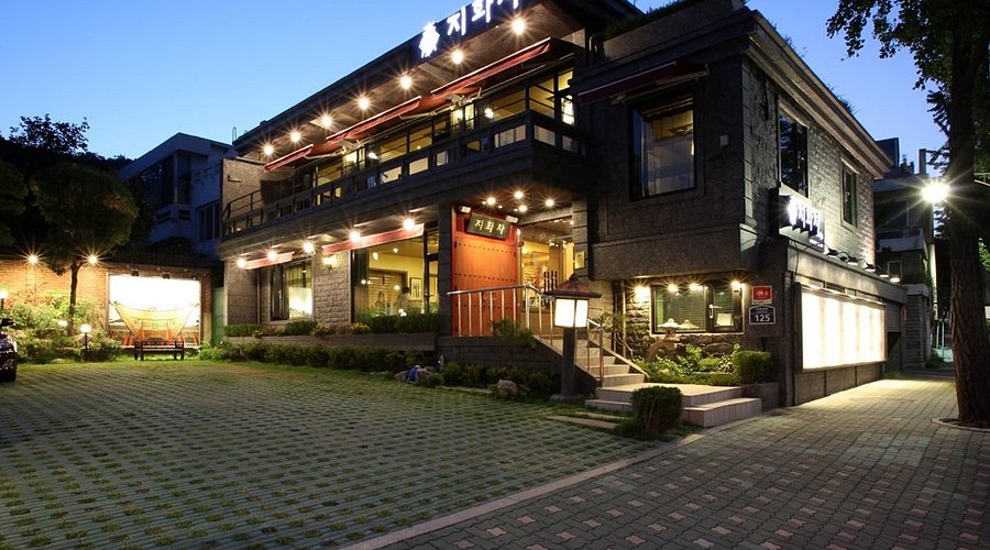 A modern, two-story restaurant with large windows, warm lighting, and a spacious, neatly landscaped front yard at dusk. The building features dark stone walls and a welcoming entrance.