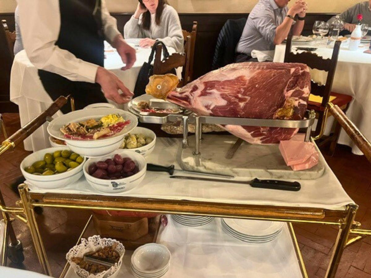 Una cameriera prepara i piatti al carrello di un ristorante con un grande prosciutto crudo, ciotole di olive, uva e antipasti di ispirazione trapanese, con i commensali seduti ai tavoli sullo sfondo.