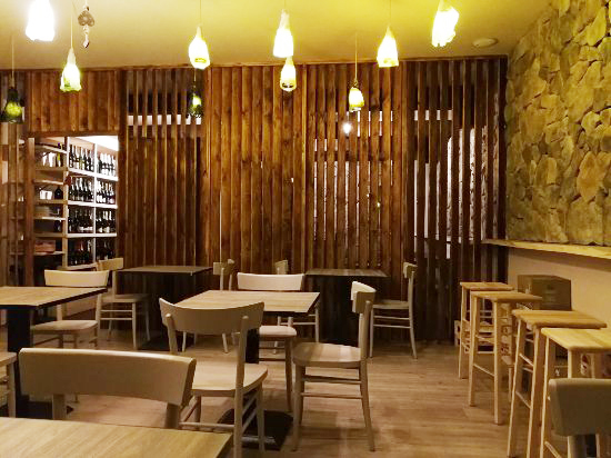 L'interno del bar è accogliente, con tavoli e sedie in legno, luci calde, una parete con doghe in legno, una parete d'accento in pietra e scaffali pieni di bottiglie sullo sfondo, che evocano il fascino di Trapani. Il caffè appare vuoto e invitante.