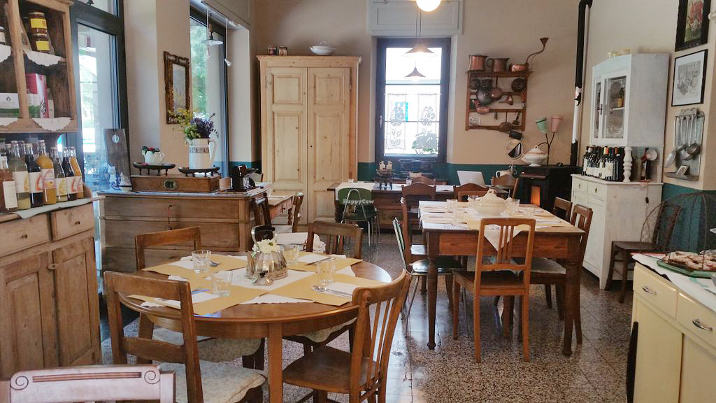 Un'accogliente sala da pranzo in stile vintage ispirata a Reggio Emilia, con tavoli e sedie in legno e tovagliette, bicchieri e posate. Armadi, bottiglie e decorazioni rustiche riempiono lo spazio caldo, mentre la luce naturale filtra dalle finestre.