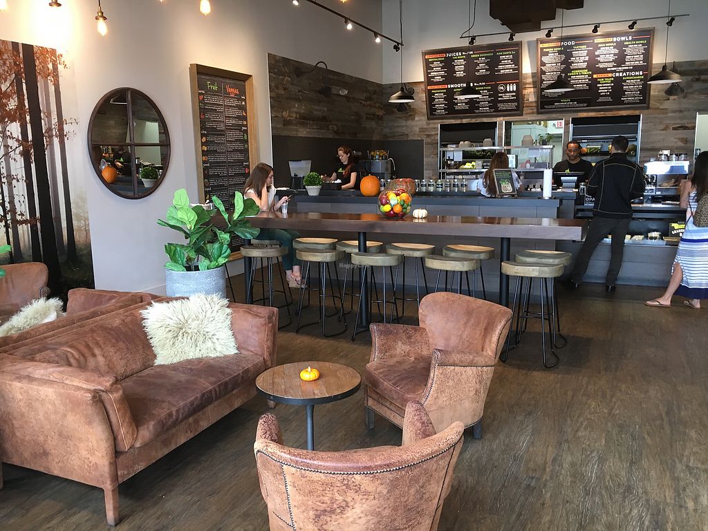 Modern cafe with cozy brown leather chairs, round tables, a bar counter with stools, plants, and autumn decorations like pumpkins. Customers are ordering at the counter, and menu boards hang on the wood-paneled wall behind.