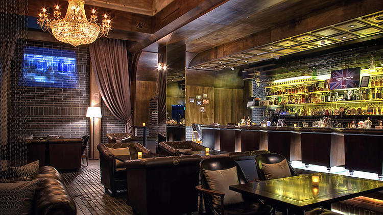 A stylish lounge bar with leather sofas, armchairs, dim lighting, a chandelier, and a long bar stocked with bottles. A TV is mounted on a brick wall, and candles are placed on tables for ambiance.