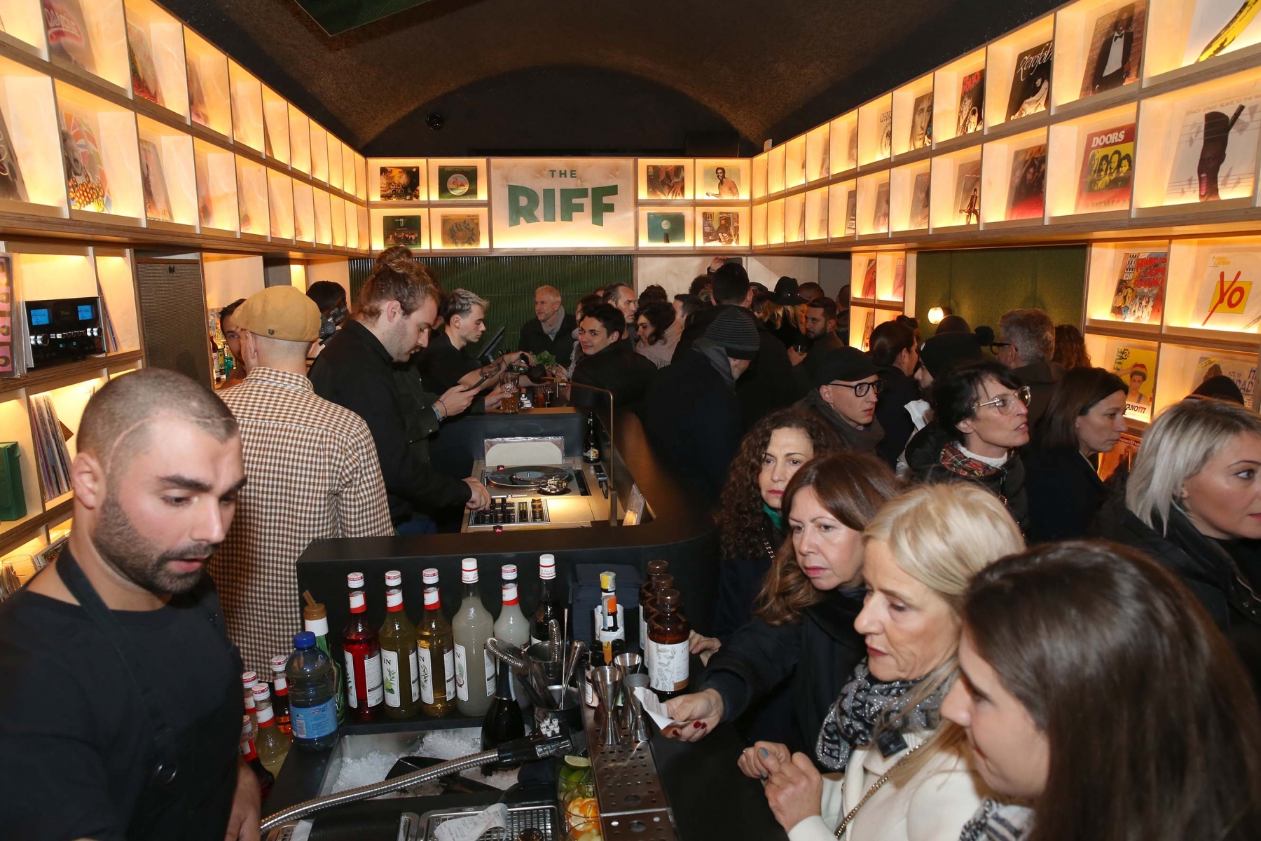 Un bar di Trapani pieno di gente che socializza. I baristi preparano i drink dietro il bancone, con bottiglie e bicchieri a vista. Lungo le pareti sono illuminati scaffali con album o libri. In fondo, un'insegna recita "THE RIFF".