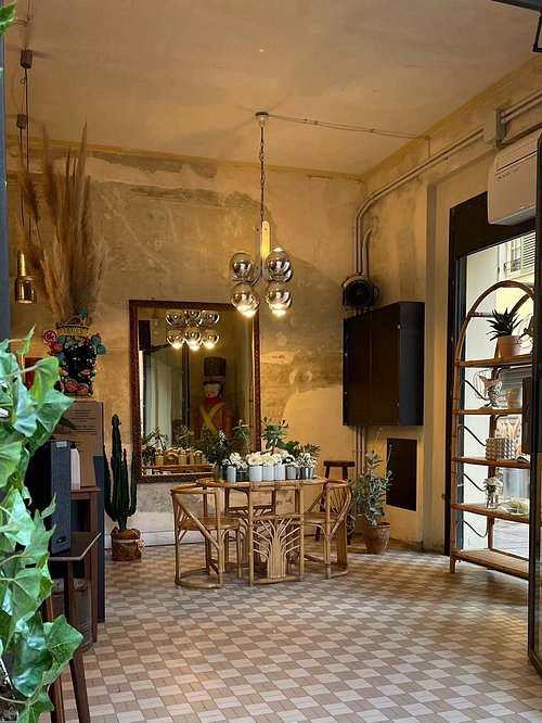 Accogliente stanza illuminata dal sole con un piccolo tavolo da pranzo in legno e sedie, circondato da piante in vaso. Un grande specchio, un lampadario, una mensola e il pavimento piastrellato conferiscono un tocco di fascino trapanese all'atmosfera calda e accogliente.