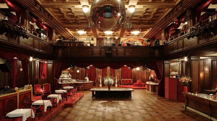 Elegante sala da ballo vintage, con pareti rivestite in legno, drappi di velluto rosso, tavoli rivestiti di bianco, una grande palla da discoteca in alto, posti a sedere sul balcone e decorazioni ornamentali per un'atmosfera lussuosa e classica.
