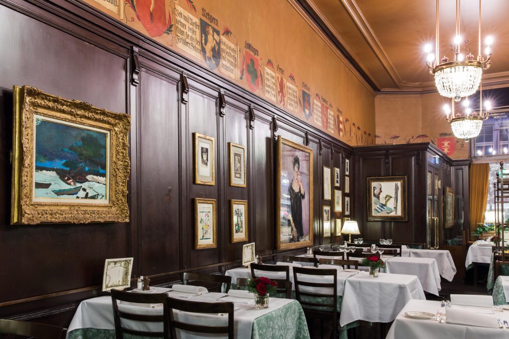 Elegante ristorante di Lugano con pareti rivestite in legno scuro, quadri incorniciati, lampadari, tovaglie bianche e tavolini apparecchiati con bicchieri e rose rosse. L'atmosfera è classica e sofisticata.