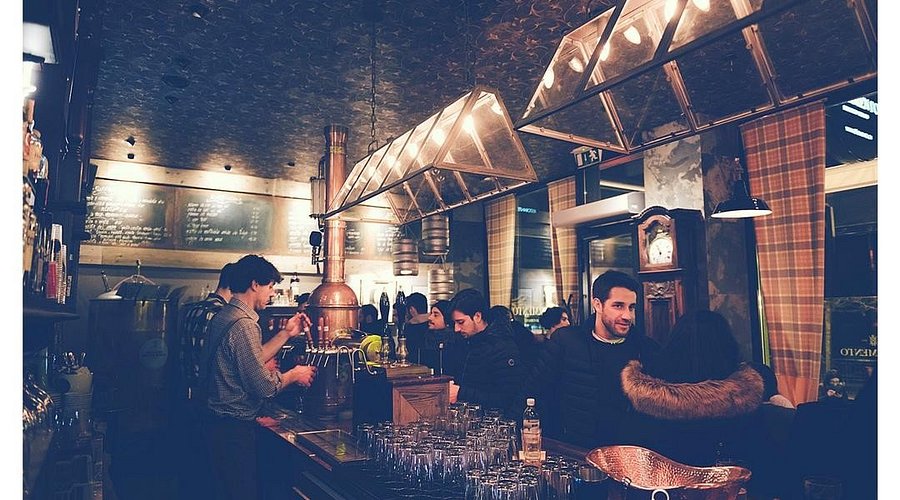 Un accogliente bar di Reggio Emilia, illuminato da una luce calda, con persone che socializzano e baristi che servono drink. I bicchieri sono allineati sul bancone e sulla parete di fondo è visibile un menu su lavagna. L'atmosfera appare rilassata e invitante.