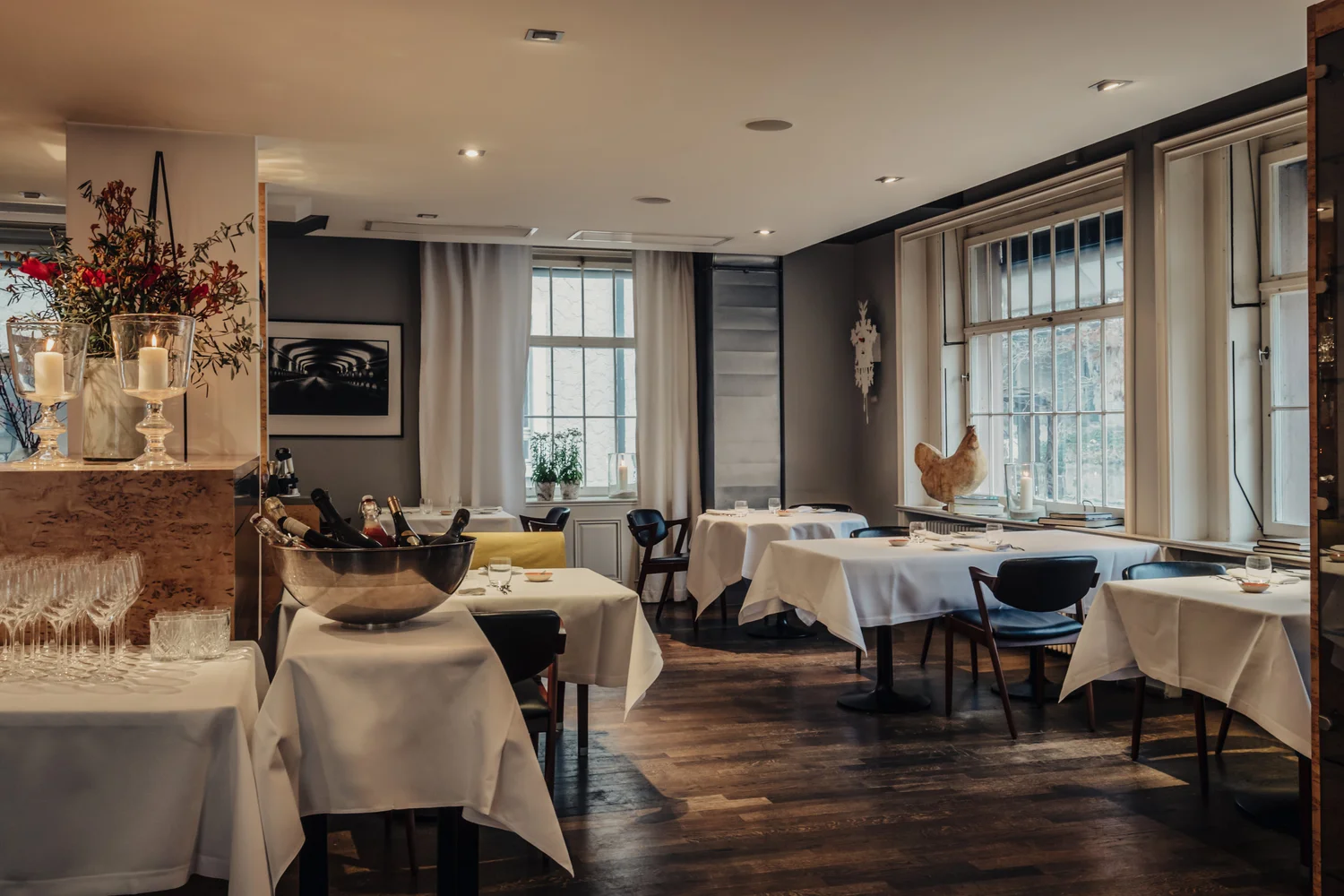 A stylish restaurant interior with neatly set tables covered in white cloths, wooden floors, modern decor, large windows, and soft lighting creating a cozy, inviting atmosphere.