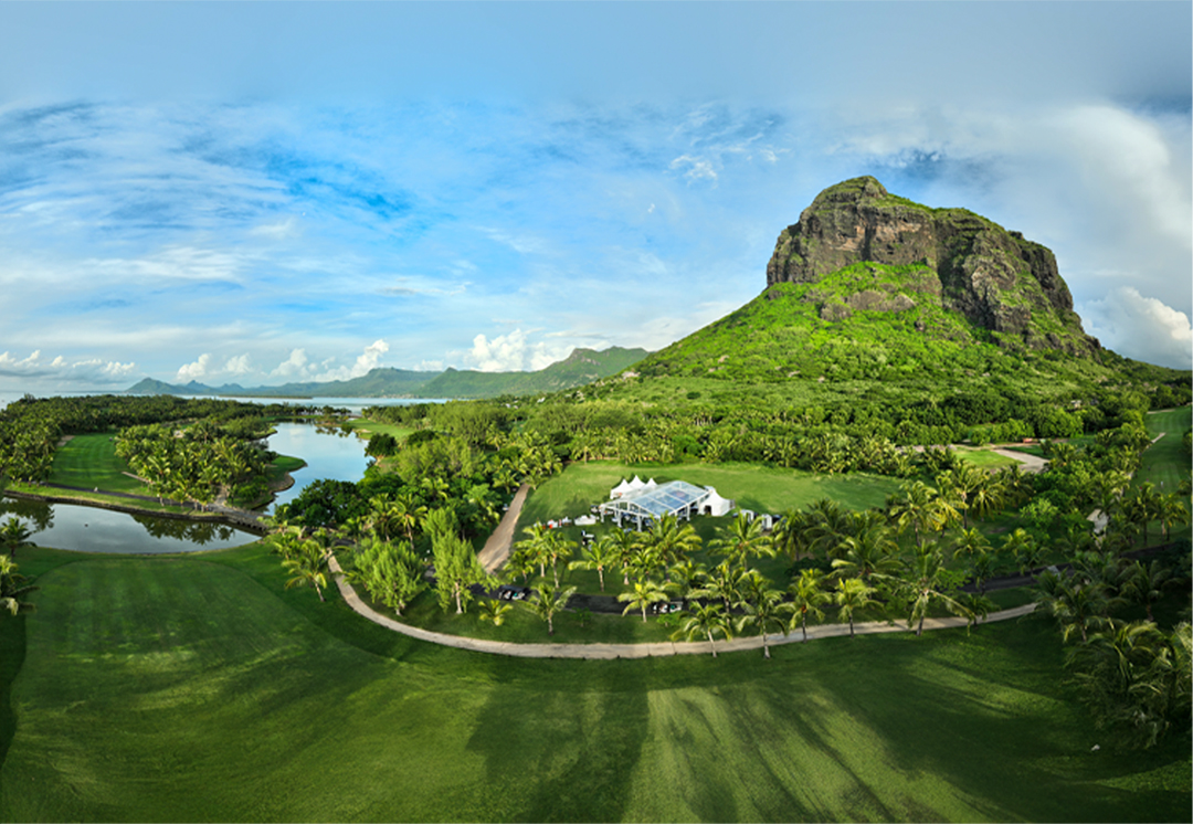 Un campo da golf verde e lussureggiante con palme, un edificio bianco, sentieri tortuosi e una grande montagna rocciosa e verde sullo sfondo, sotto un cielo blu con nuvole sparse.