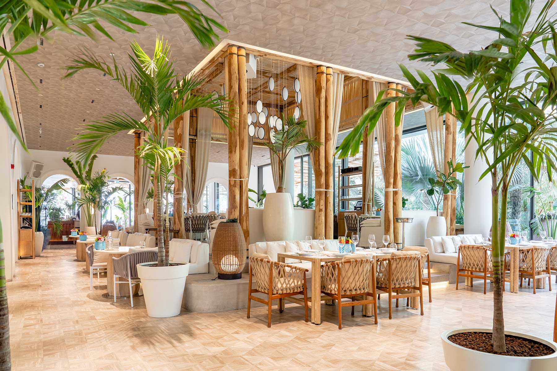 A spacious, modern restaurant with natural light, wooden furniture, white cushions, potted palm plants, and bamboo columns, creating a tropical and elegant atmosphere with neatly set dining tables.