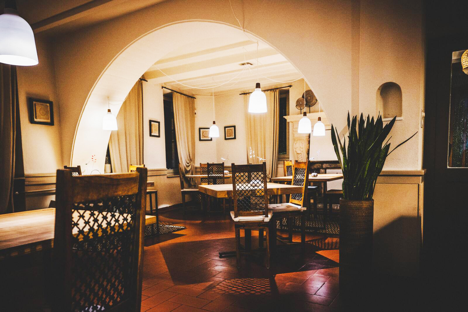 L'interno di un ristorante di Reggio Emilia, accogliente e illuminato da luci calde, con tavoli e sedie in legno, una porta ad arco, una pianta in vaso, opere d'arte incorniciate sulle pareti color crema e luci a sospensione che creano un'atmosfera invitante.