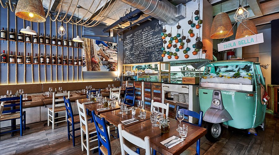Un ristorante accogliente con tavoli rustici in legno, sedie bianche e blu, luci appese, una parete di bottiglie di vino, un menu su lavagna e un motorino verde vintage trasformato in un'esposizione di frutti di mare.