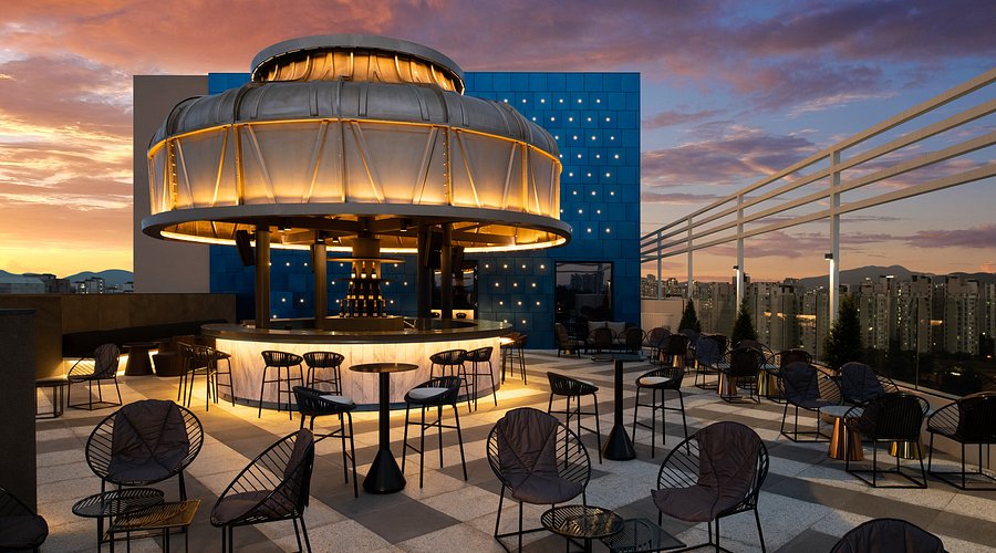 Rooftop bar at sunset with a circular, illuminated canopy over the bar area, surrounded by modern black chairs and tables, city buildings visible in the background under a colorful sky.