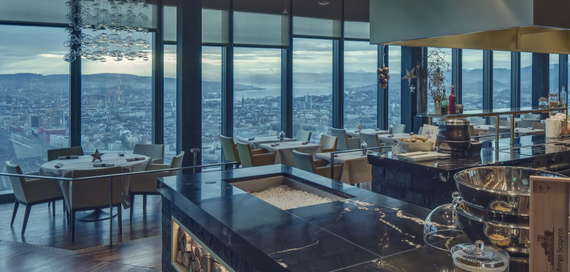 Un moderno ed elegante ristorante di Lugano è caratterizzato da vetrate che si affacciano sul paesaggio urbano e sulle colline lontane al tramonto. I tavoli sono apparecchiati per cenare, con un elegante bancone di marmo nero in primo piano.
