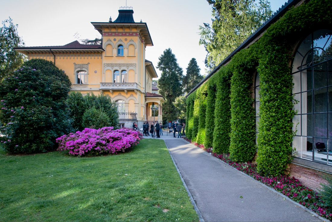 Una villa gialla dall'architettura ornata sorge accanto a un giardino lussureggiante, con fiori rosa sbocciati che evocano il fascino di Reggio Emilia. Un sentiero lastricato corre tra la casa e un edificio ricoperto di edera, con persone che passeggiano sullo sfondo.